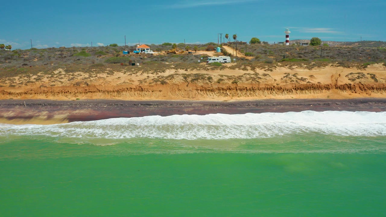 Opening aerial frame of the San Jose Del Cabo coast with green ocean in front, white surf on a dark wet-sand strip, ochre bluff, scattered buildings, palms, and no visible people.