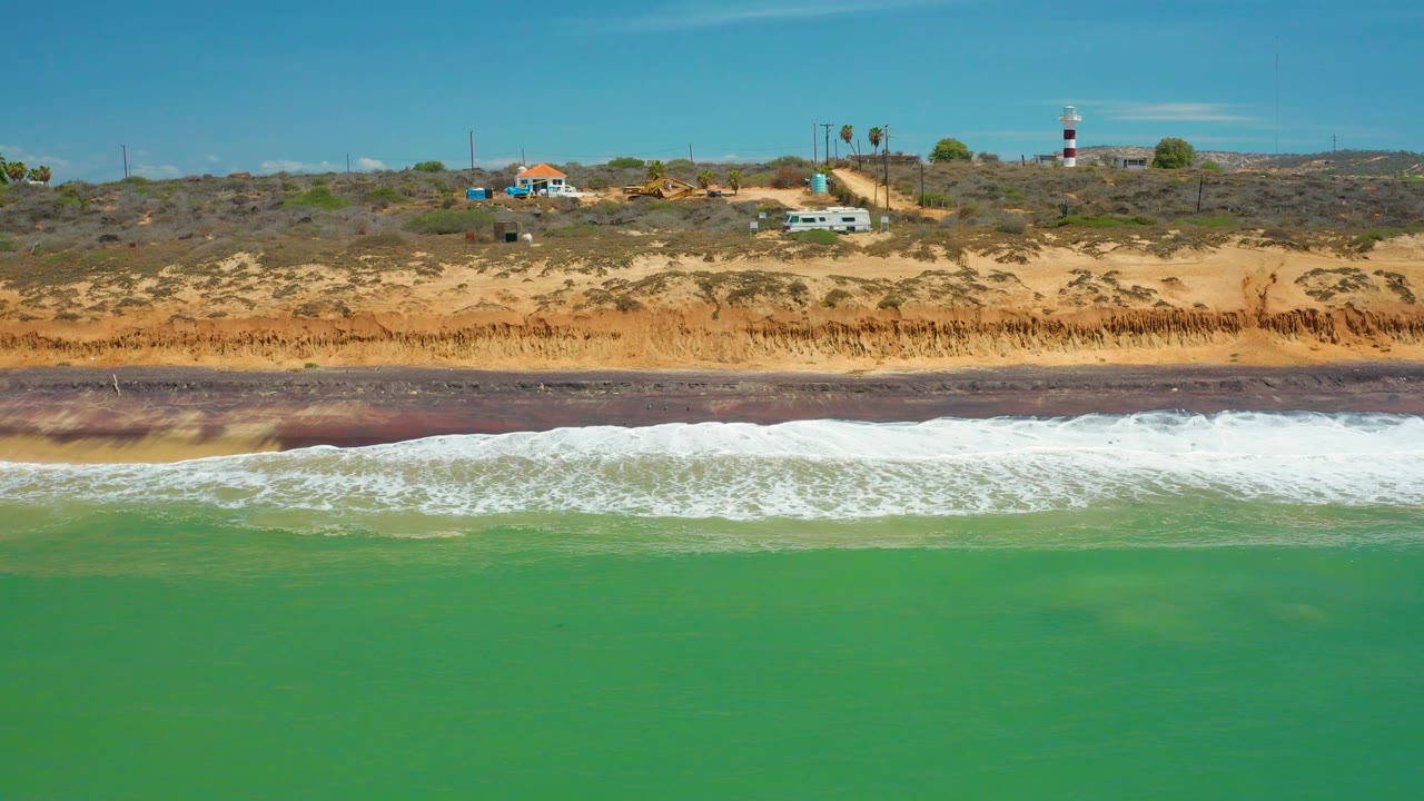 San Jose Del Cabo shoreline frame with white waves spread across the lower beach, reddish dark wet sand behind the foam, ochre bluffs with brush, cliff-top buildings, and no visible people.