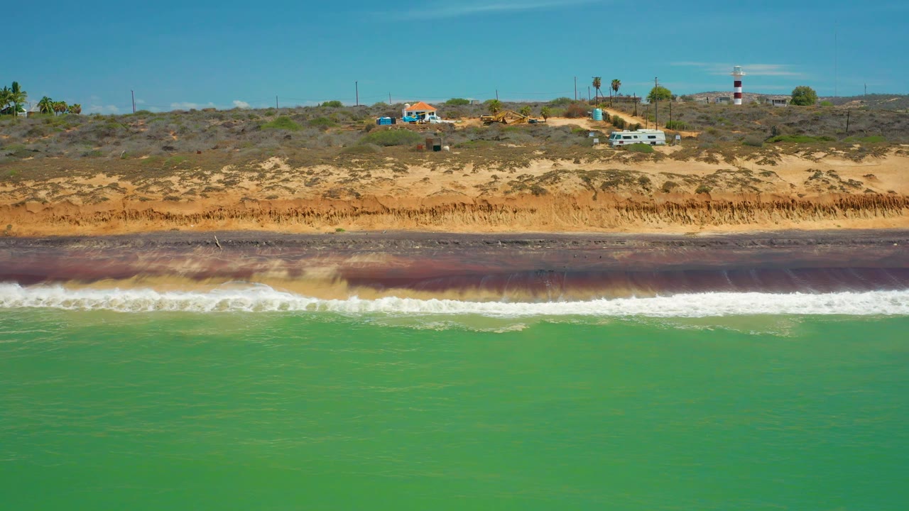 Aerial coastal frame from San Jose Del Cabo, Mexico, showing green water, foamy white surf, an orange sandy bluff, scattered buildings, palms, a striped tower, and no visible people.