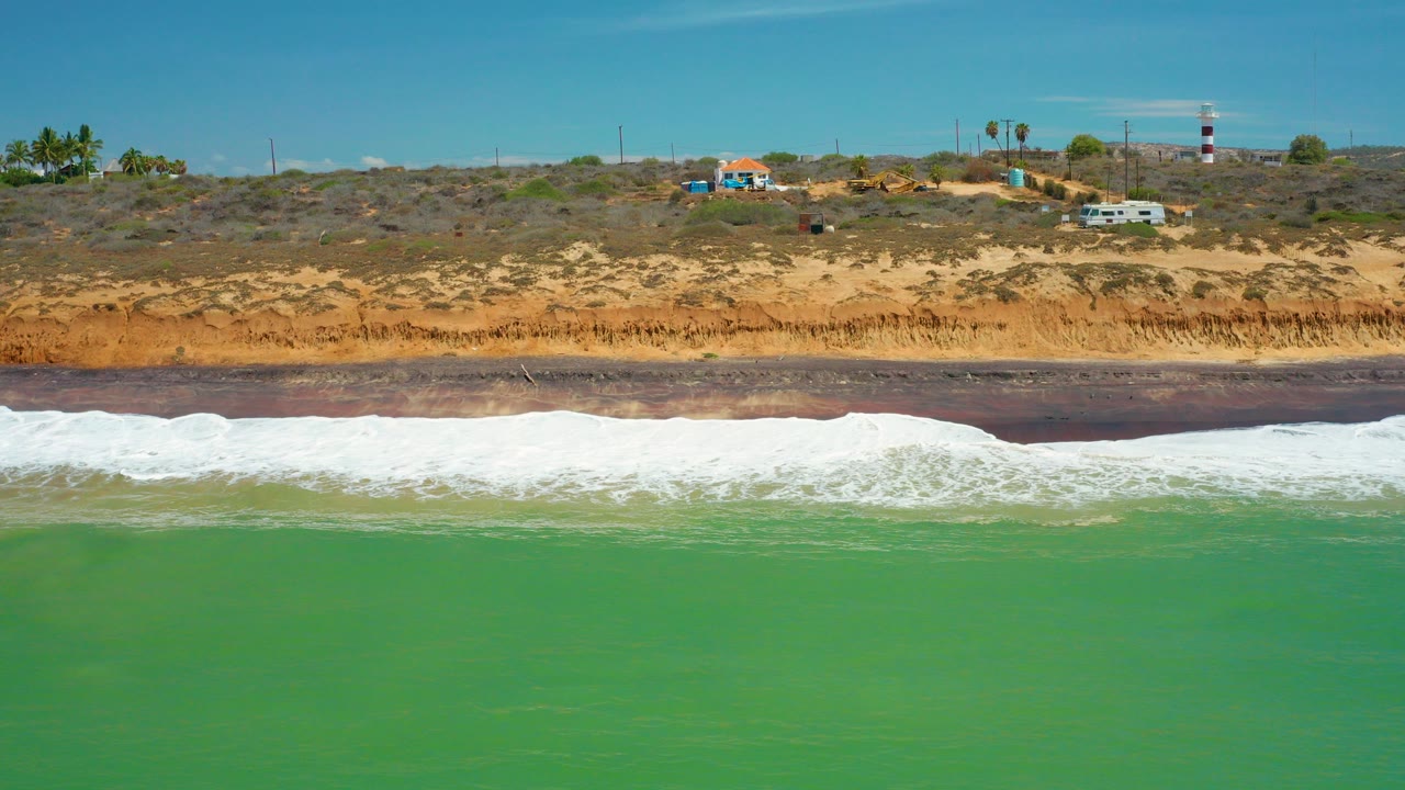 At 0:00, green water and white surf reach a dark sand beach in San Jose Del Cabo, Mexico, with a dry bluff, small buildings, vehicles, palms, and a striped tower beyond, no visible people.