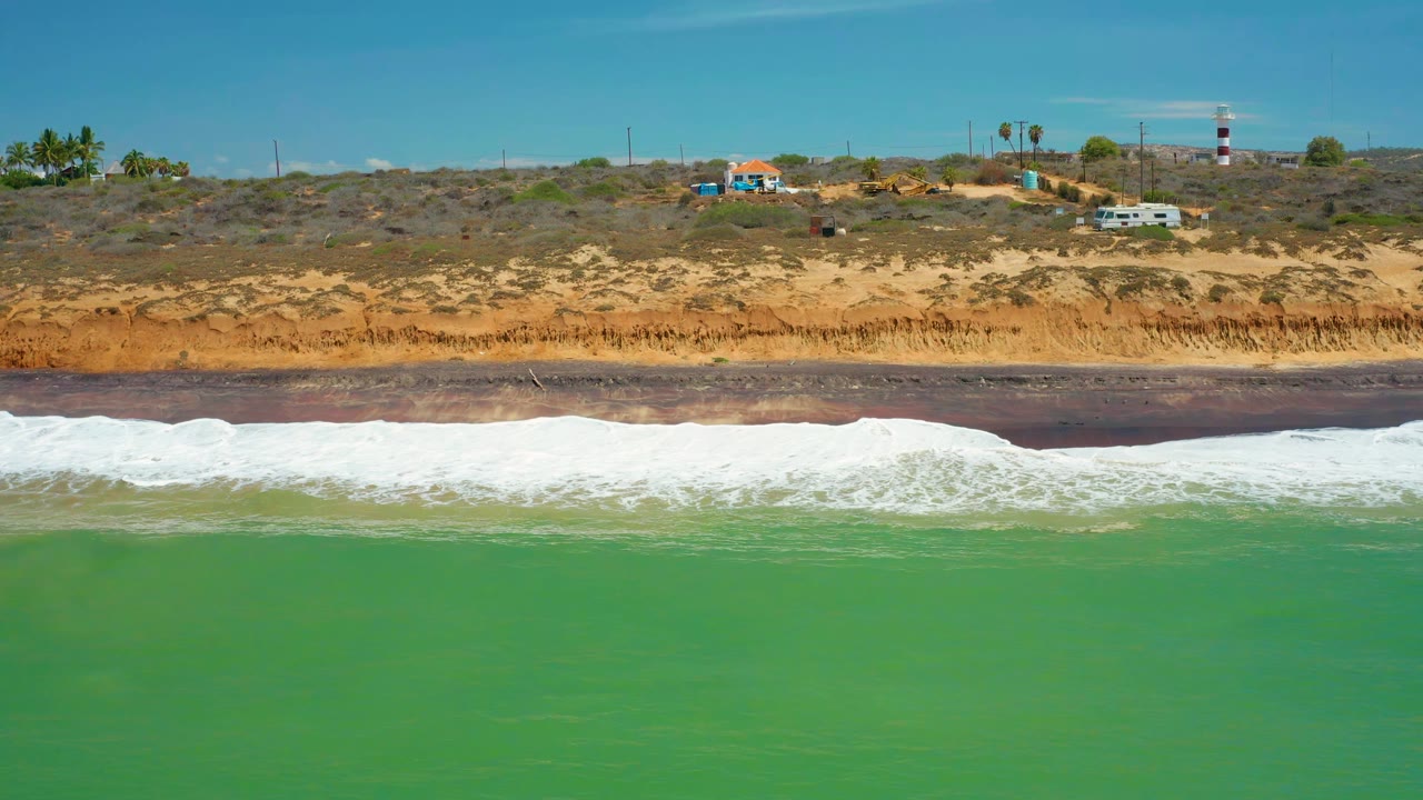 At 0:01, a wide white foam band spreads along the dark shoreline in San Jose Del Cabo, Mexico, while low vegetation, small buildings, vehicles, palms, utility poles, and a striped tower line the bluff, no visible people.