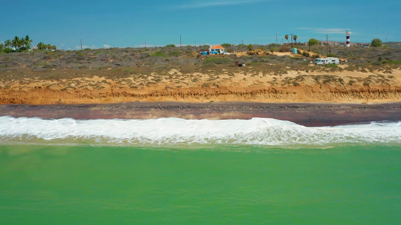At 0:02, the foam line appears thicker along the beach in San Jose Del Cabo, Mexico, with green water in front and a bluff topped by scattered structures, vehicles, palms, utility poles, and a striped tower, no visible people.