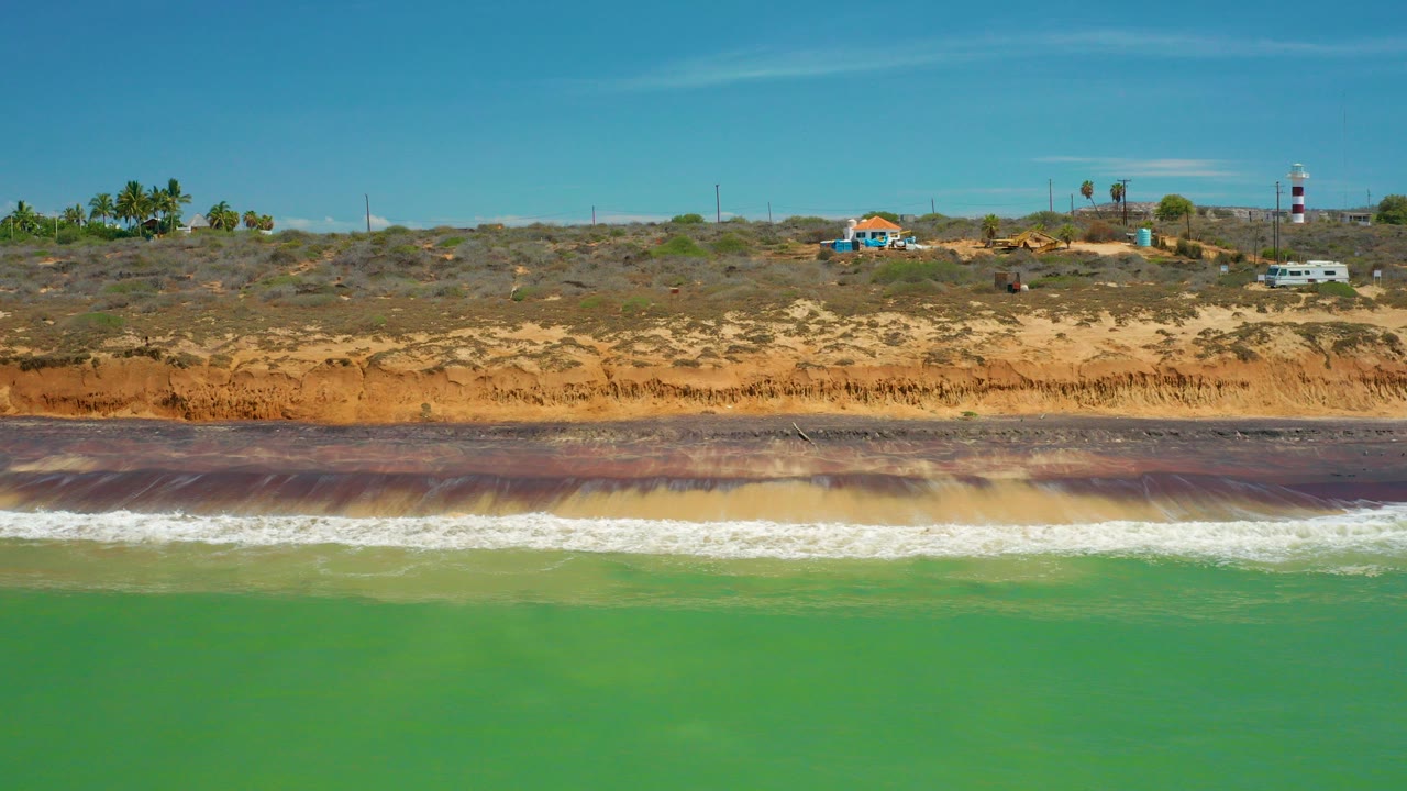 Green water breaks into a white surf line below brown bluffs and sparse coastal buildings in San Jose Del Cabo, Mexico, with no visible people.