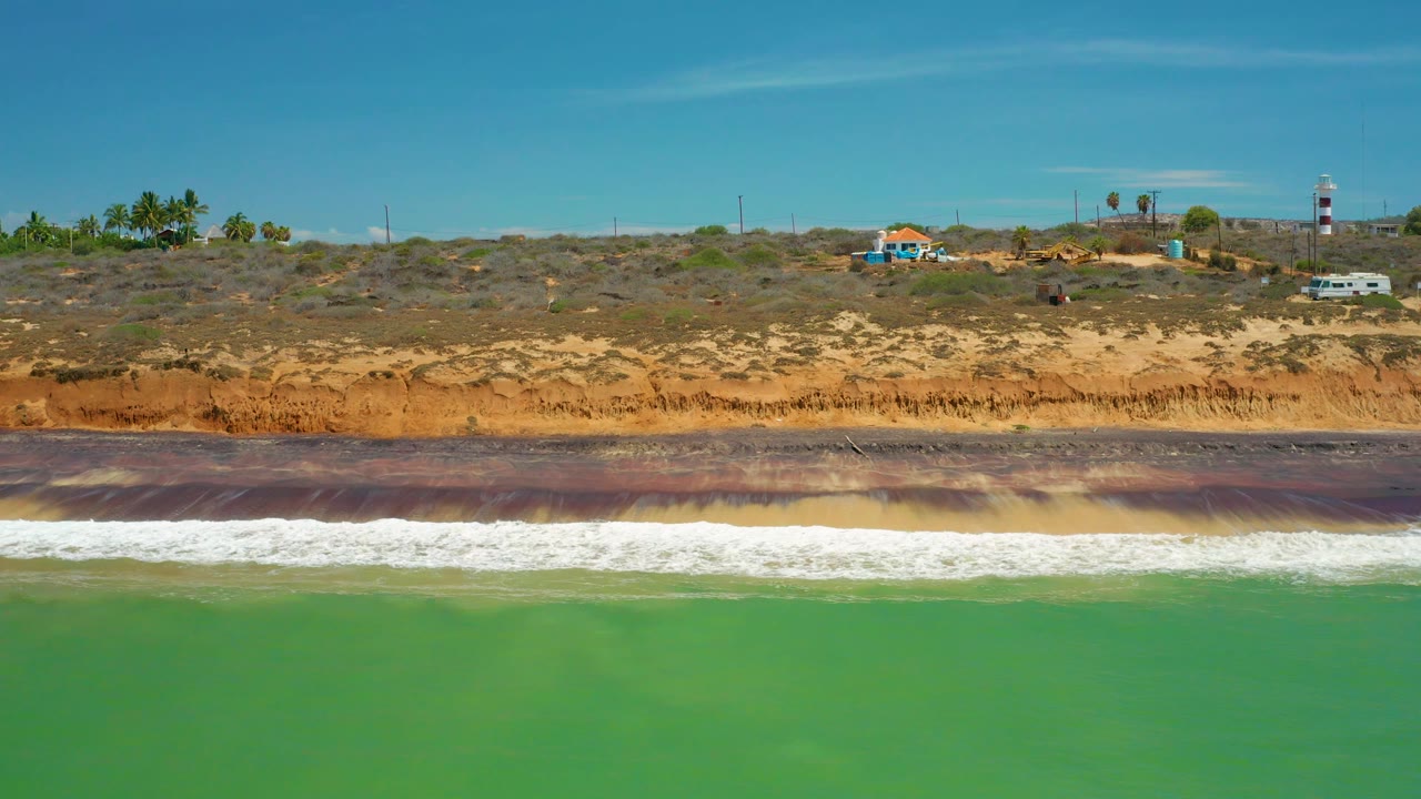 A broad empty-looking San Jose Del Cabo shoreline holds green water in front, a white breaking wave line, brown bluffs, scattered palms, and distant structures, with no visible people.
