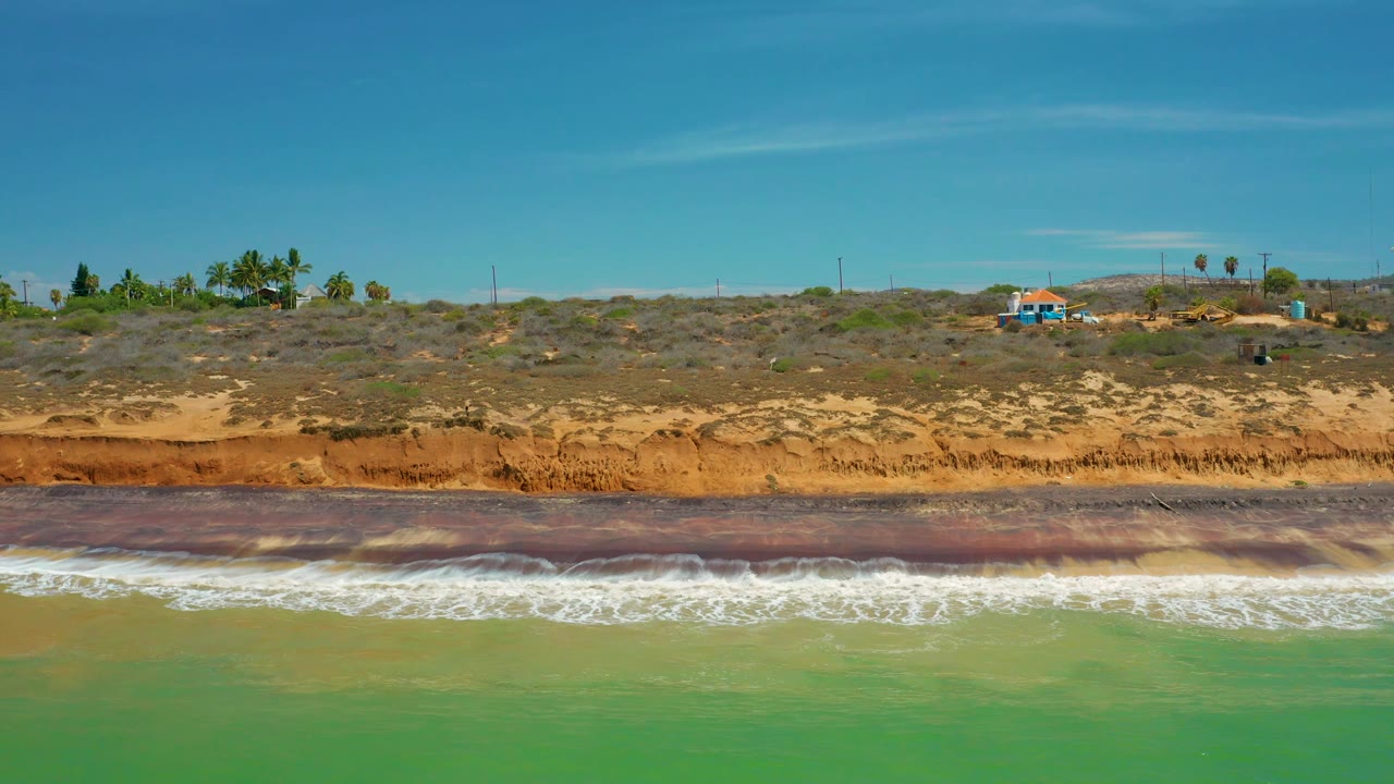 San Jose del Cabo coastal frame showing surf breaking across green water, a reddish beach shelf, scrub-covered dunes, utility poles, palms, and a small blue-roofed building inland.