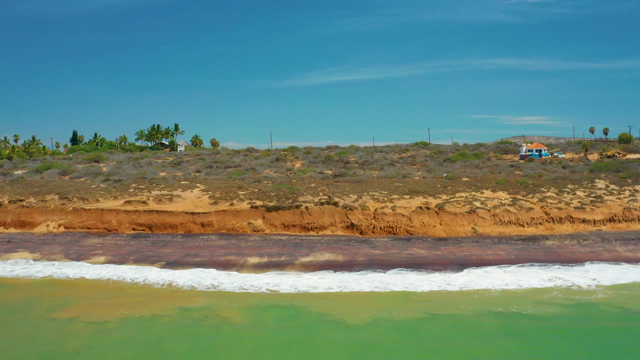 San Jose del Cabo coastline with green water in the foreground, a white surf band, an orange eroded bluff, scrub vegetation, small buildings, and no visible people.