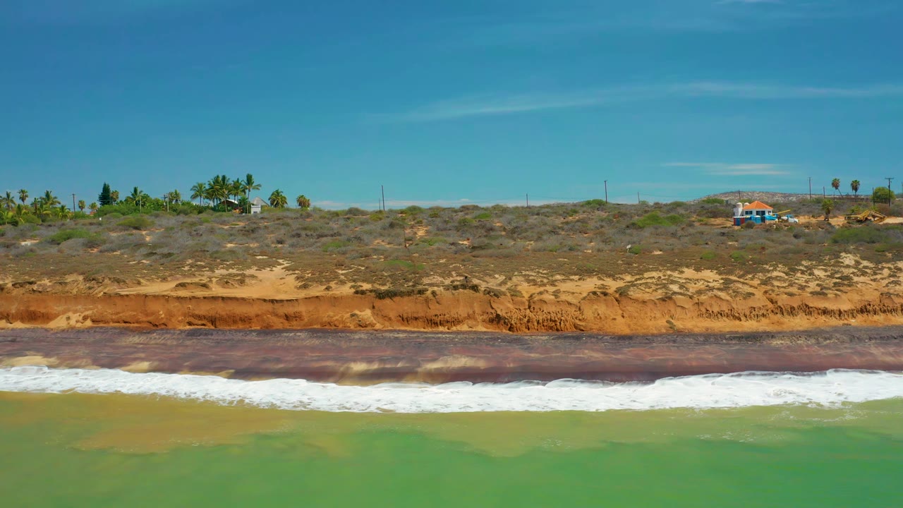San Jose del Cabo shoreline with whitewater spreading across green water, the brownish orange bluff centered behind it, and no visible people on the land or beach.