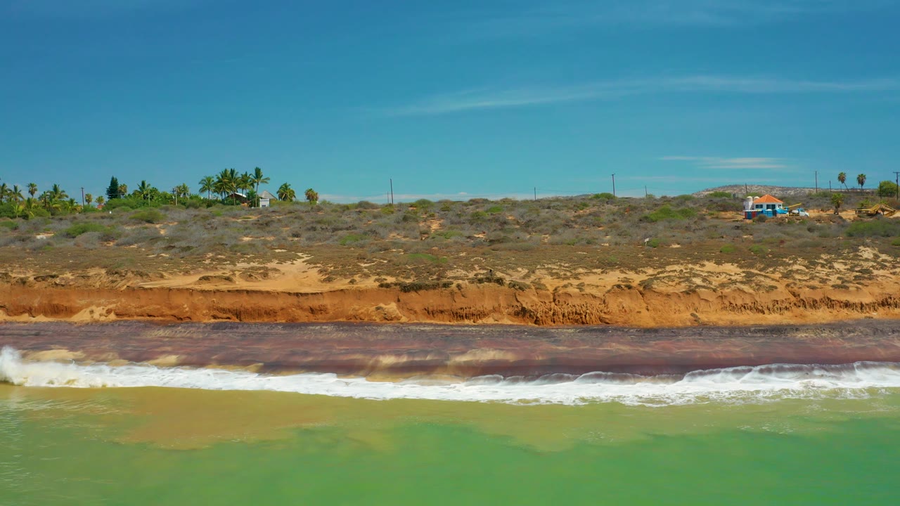 San Jose del Cabo coastal bluff with green sea in front, white waves along the base, scrub plants across the top, distant poles, and no visible people.