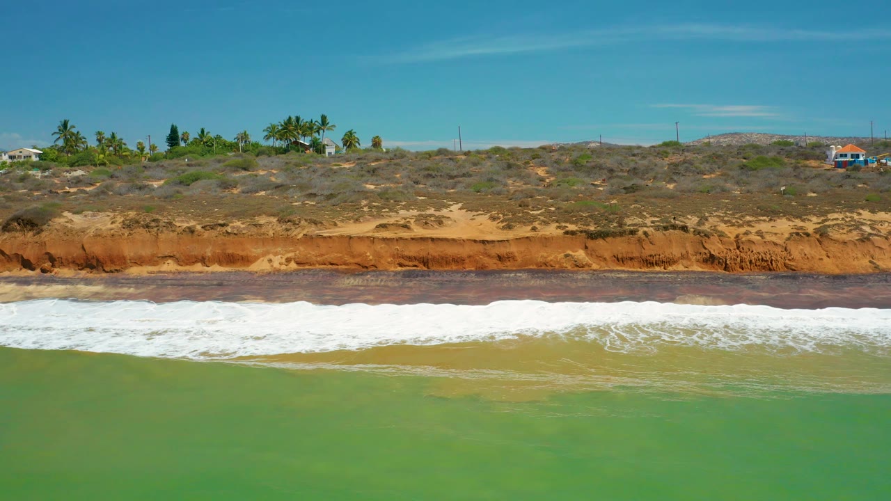 San Jose Del Cabo shoreline with foamy white water stretched along the beach, green sea in front, palms and scrub on the bluff, distant structures, and no visible people.