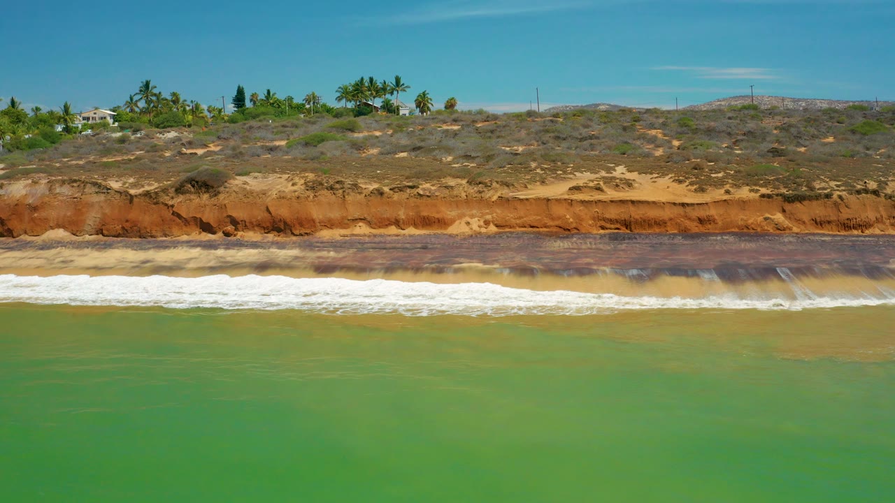A breaking wave crosses the middle of the San Jose del Cabo frame, with green water below, scrub-covered bluffs above, small distant buildings near palms, and no visible people.
