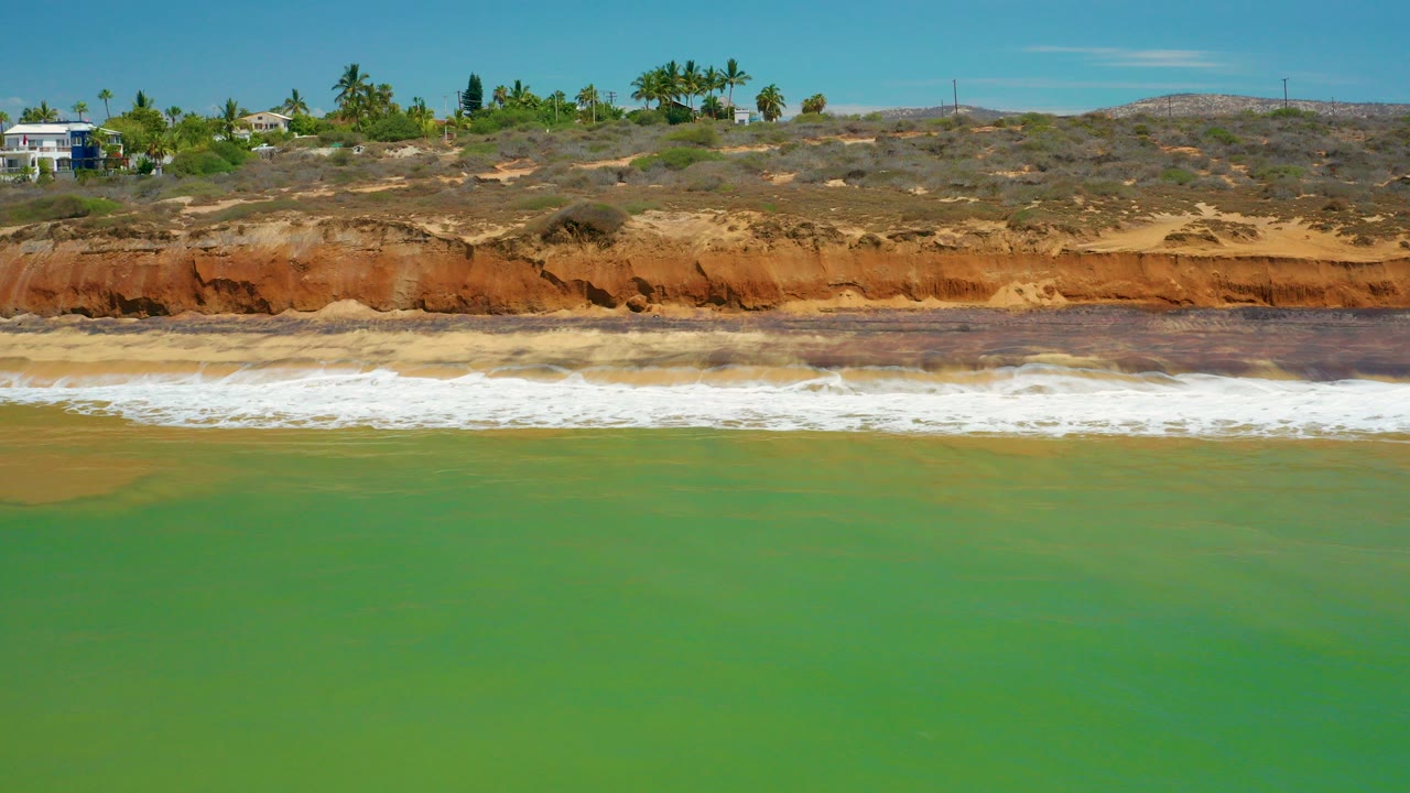 Opening frame of the San Jose Del Cabo shoreline with green sea in the foreground, white surf at the sand, red-orange bluff above the beach, palm trees, a white building on the left ridge, and no visible people.