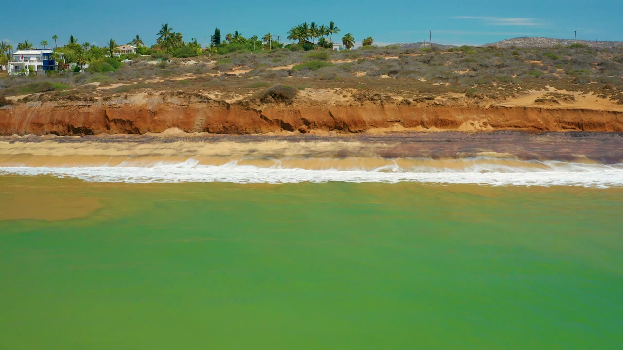 At 0:02, white waves spread across the beach below the red bluff, with green water in front and several blufftop structures and palms visible along the San Jose Del Cabo ridge, with no visible people.