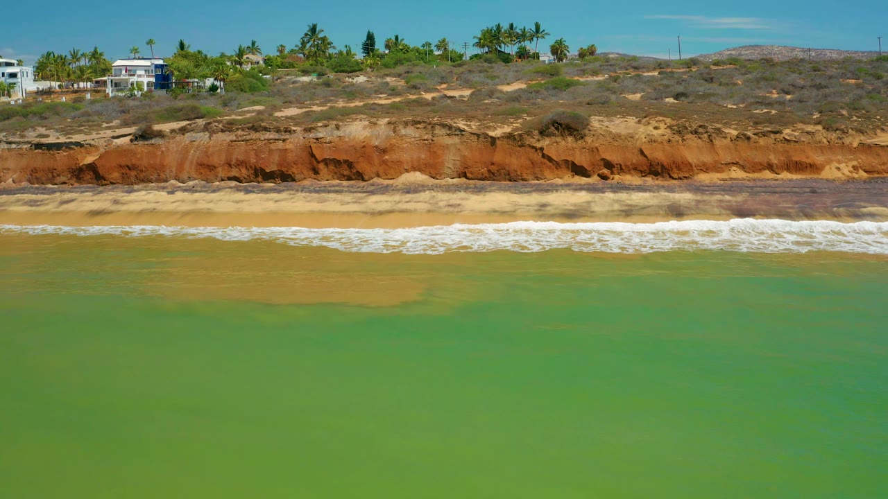Green water and a foamy surf band sit below eroded reddish cliffs, with palms and low buildings along the ridge in San Jose del Cabo, Mexico, with no visible people.