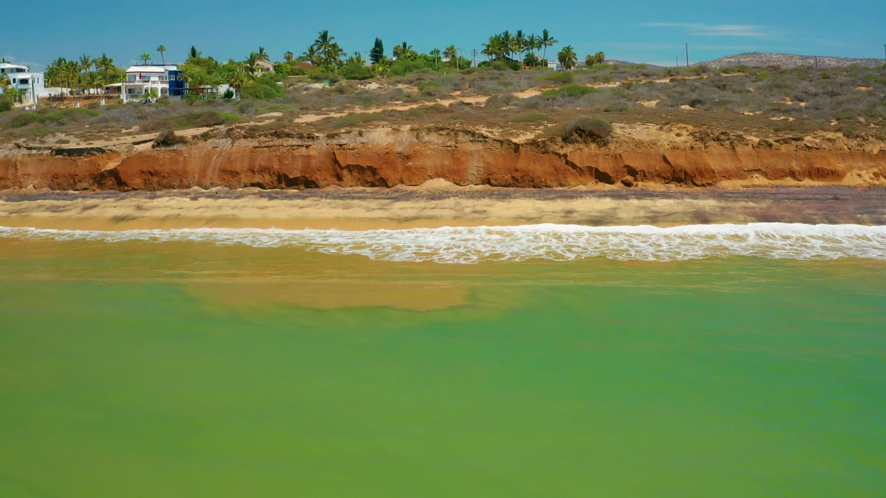 A wide view holds green ocean in front, a pale surf line, rust and tan coastal bluffs, palms, and several hillside homes in San Jose del Cabo, Mexico, with no visible people.