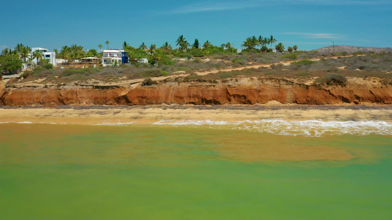 At 0:00 in San Jose Del Cabo, Mexico, shallow green-blue water leads to a sandy beach, orange bluff, hillside homes, palms, and blue sky, with no visible people.