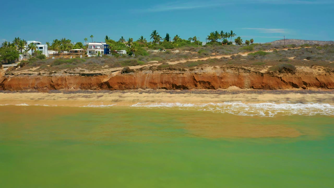 At 0:01 in San Jose Del Cabo, Mexico, a thin wave breaks along the sand below orange bluffs, with white homes and palms on the ridge and no visible people.