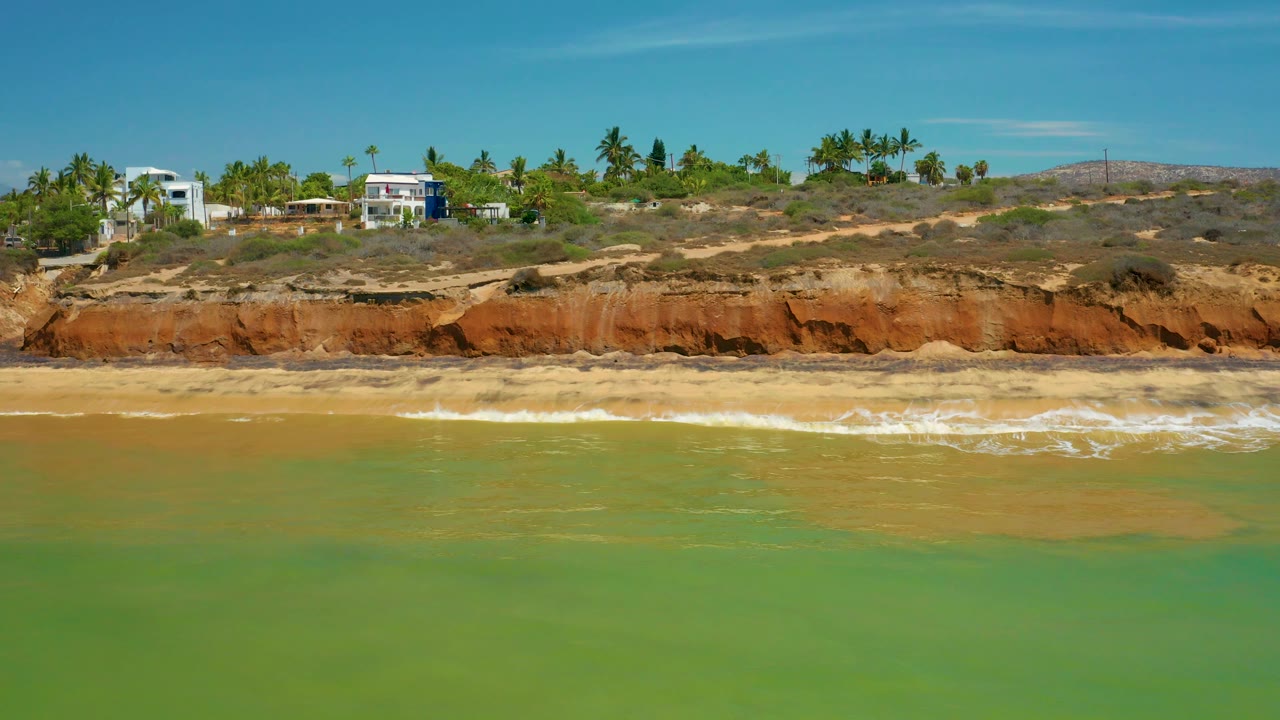 At 0:02 in San Jose Del Cabo, Mexico, the shoreline spans the frame with green water in front, white foam on sand, orange cliffs, palms, hillside houses, and no visible people.