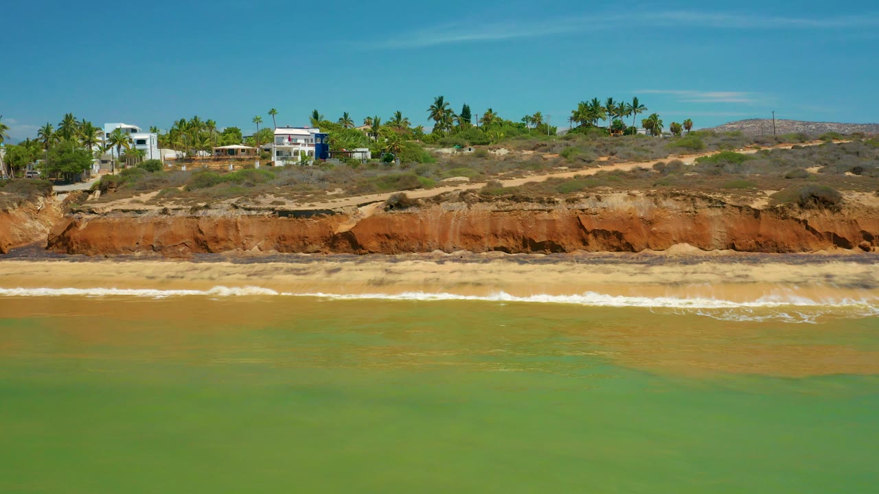 Beach surf runs below a long orange bluff with homes and palms above.