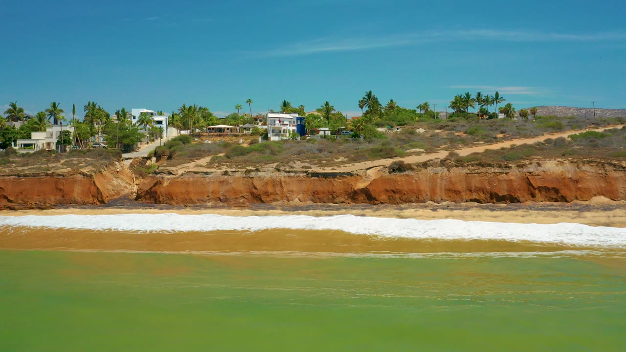 San Jose Del Cabo shoreline with green water in front, a white wave band on the beach, red rock bluff, palm trees, and homes on the ridge, with no visible people.