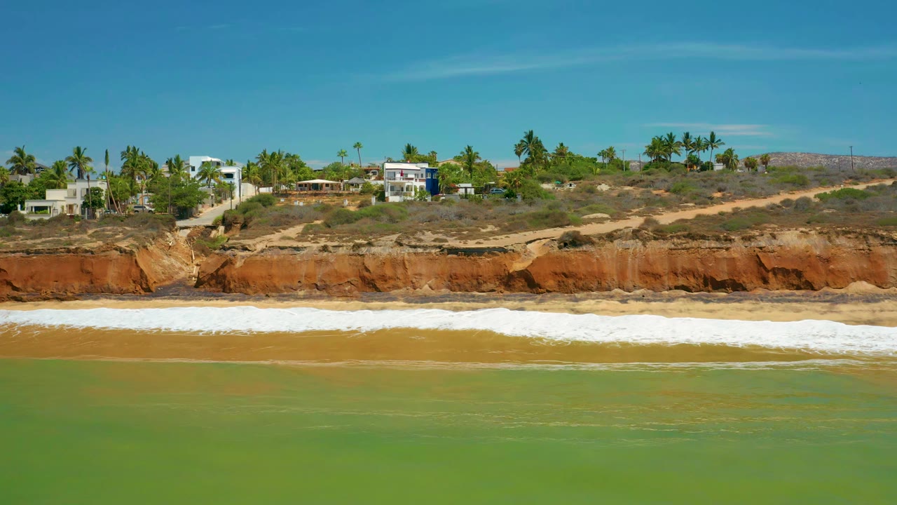 A slightly later view of the San Jose Del Cabo coast shows the white surf line stretched below reddish cliffs, with homes and palm trees along the upper edge and no visible people.