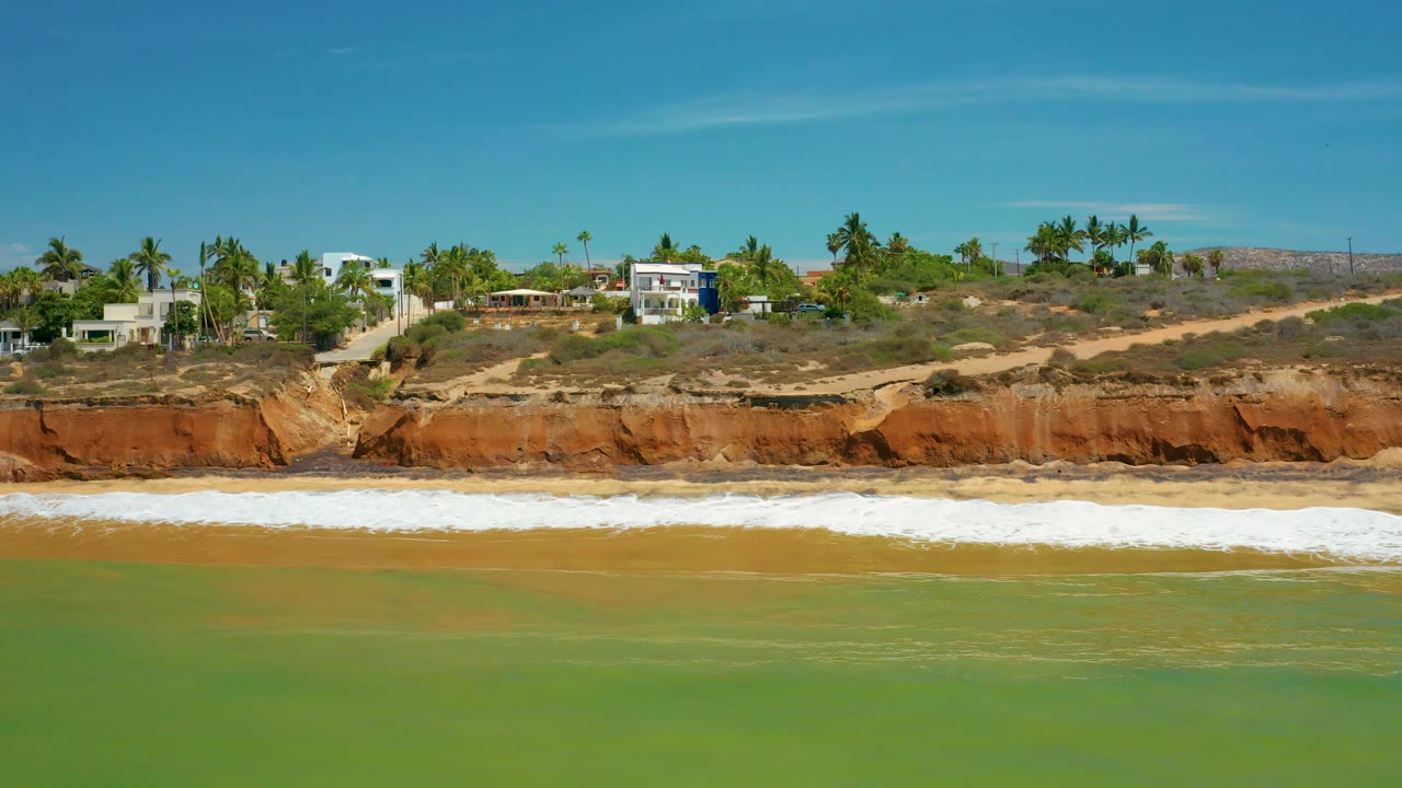 San Jose Del Cabo beach view with muted green water in the foreground, a bright breaking wave near shore, red eroded bluff behind it, and residential buildings above, with no visible people.