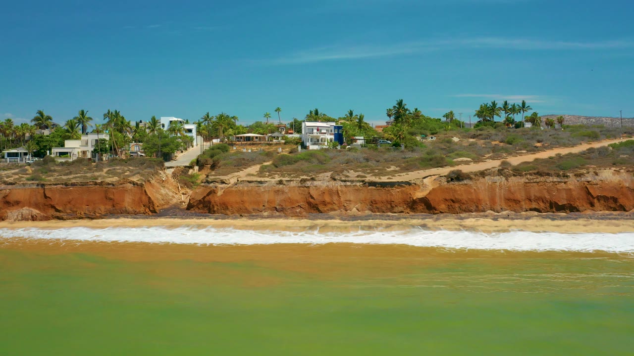 Coastal houses and palms above a red bluff with white surf below.
