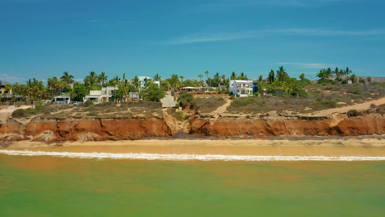 Wide 4K view of San Jose Del Cabo coastline with green-blue ocean in front, white surf at the beach, red bluff rock, palm trees, and houses above the shore, no visible people.