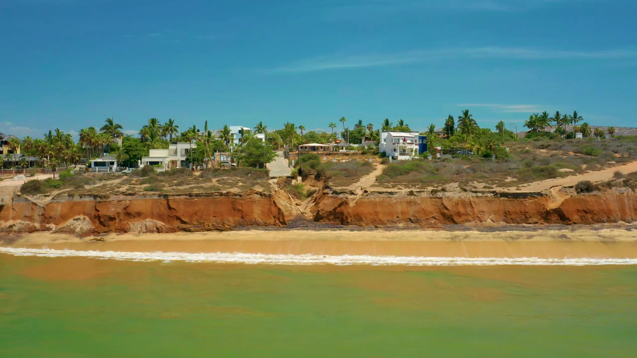 San Jose Del Cabo surf breaks along a narrow beach below red eroded cliffs, with palm trees and light-colored houses spread across the bluff, no visible people.