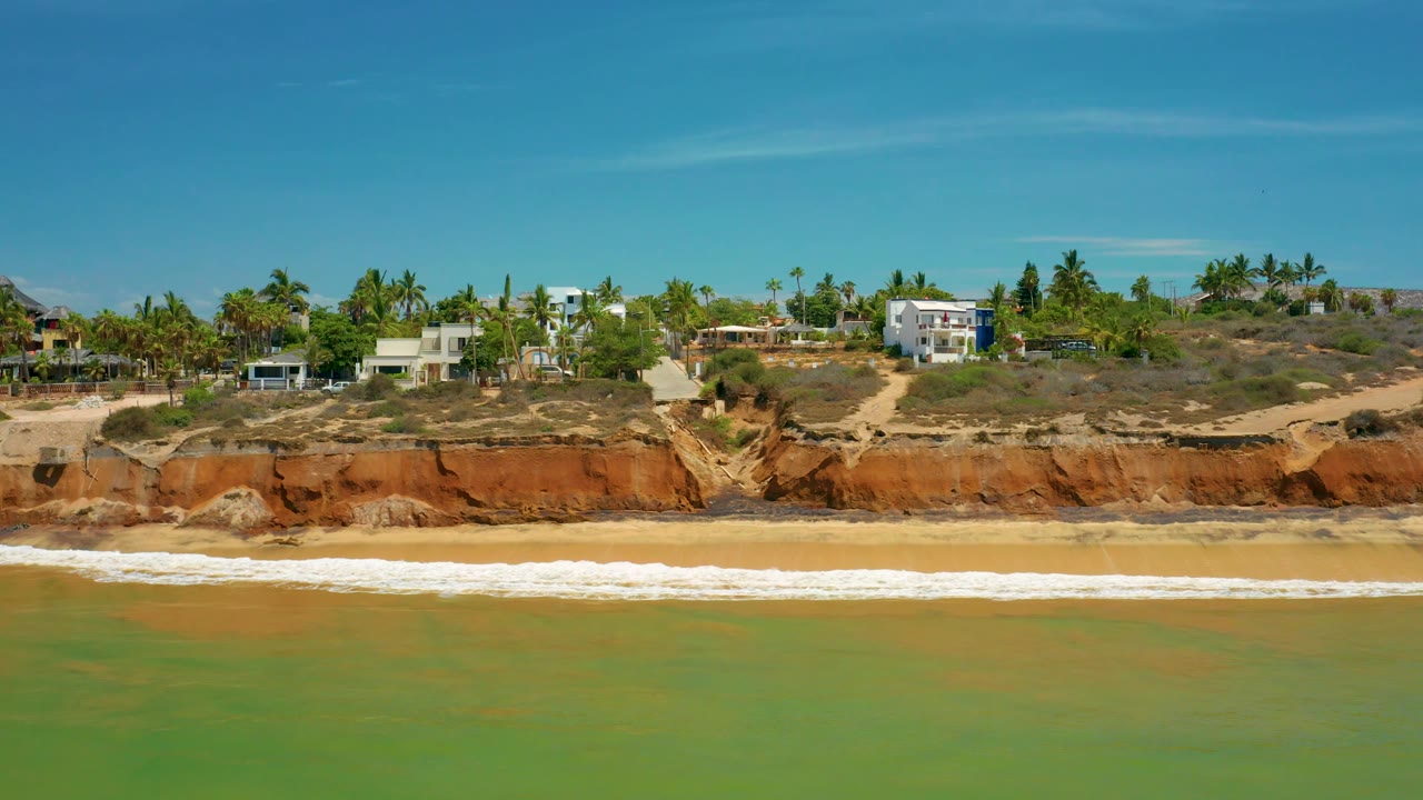 A wide aerial-looking frame shows waves running parallel to a rocky San Jose Del Cabo beach, with red bluff faces, palms, sandy tracks, and houses in bright daylight, no visible people.