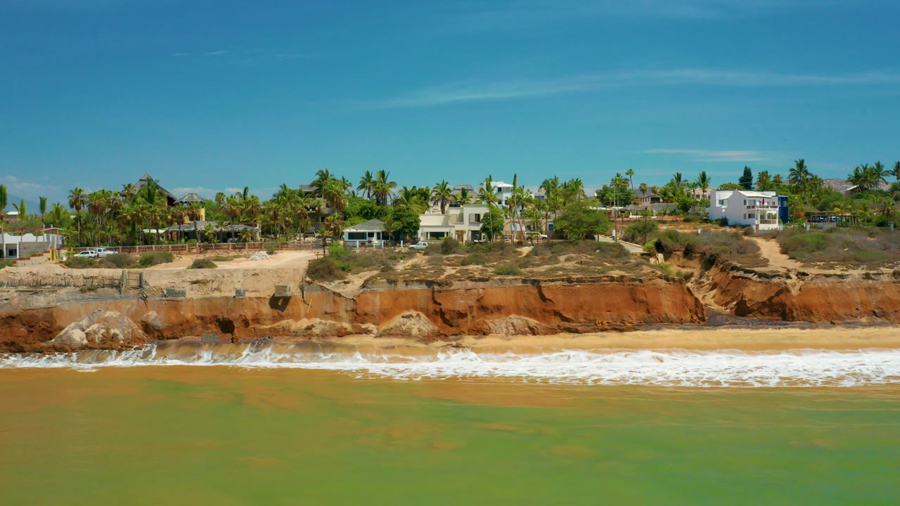 Opening aerial view of the San Jose del Cabo shoreline with green-blue water in front, white surf on the sand, red cliffs, homes, palm trees, and no visible people.