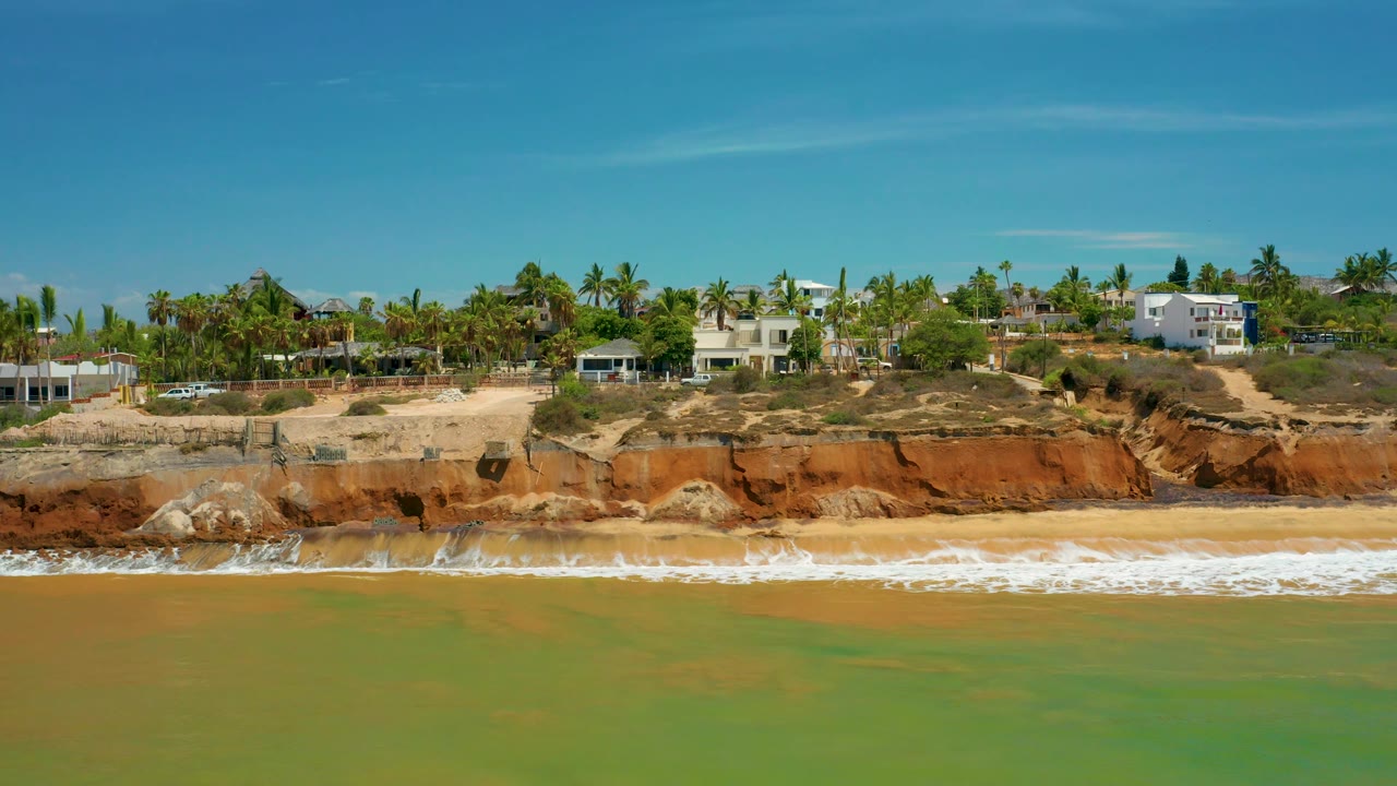 San Jose del Cabo aerial frame with mottled green water in the foreground, a narrow surf band, red cliff face, and low white buildings among palms with no visible people.
