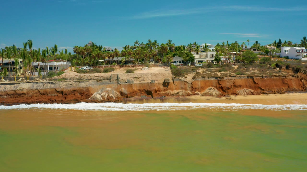Aerial view of green sea water approaching a sandy San Jose Del Cabo beach below orange rock cliffs, palms, and white buildings, with no visible people.