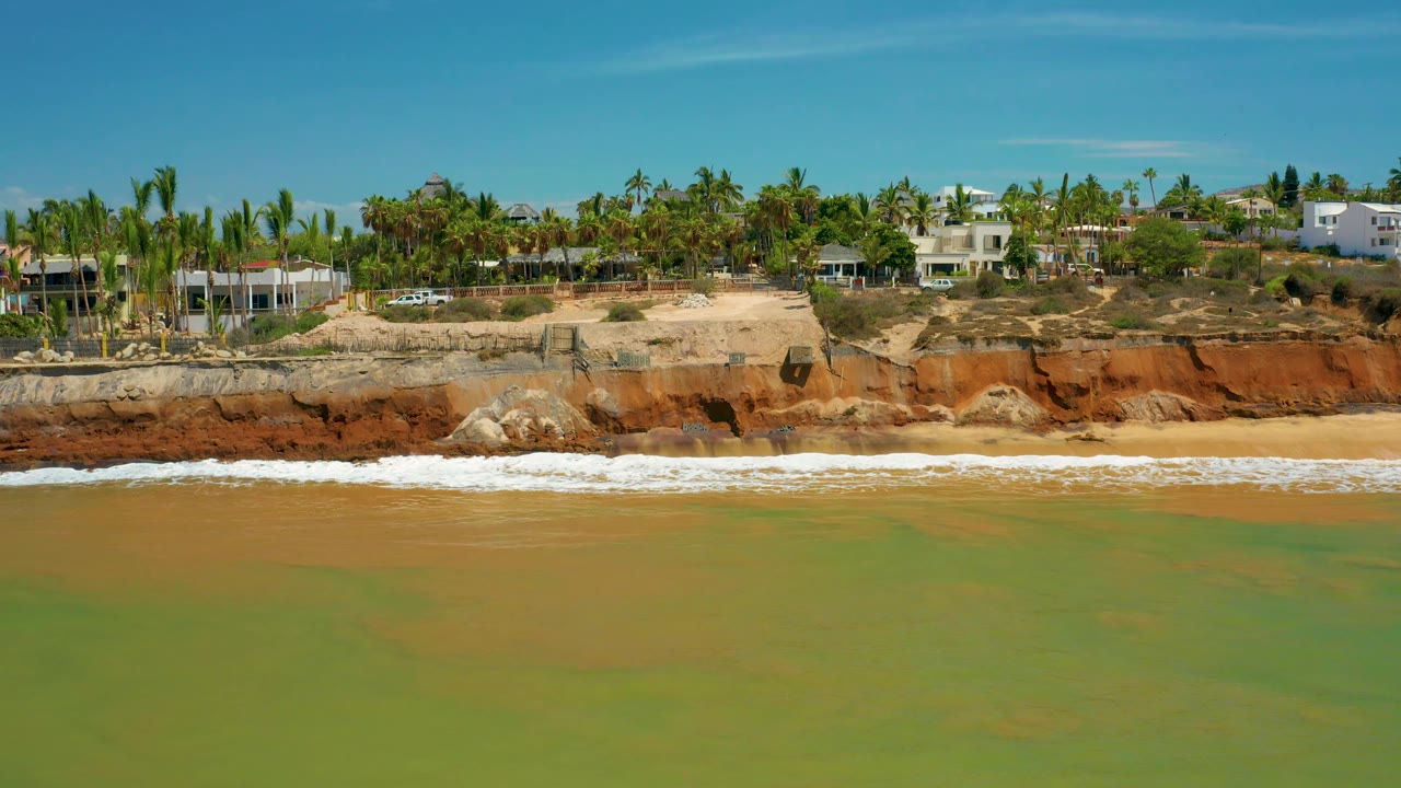 A broad aerial frame shows San Jose Del Cabo green water, a white band of wash on the sand, orange cliff layers, palms, and low-rise residences, with no visible people.