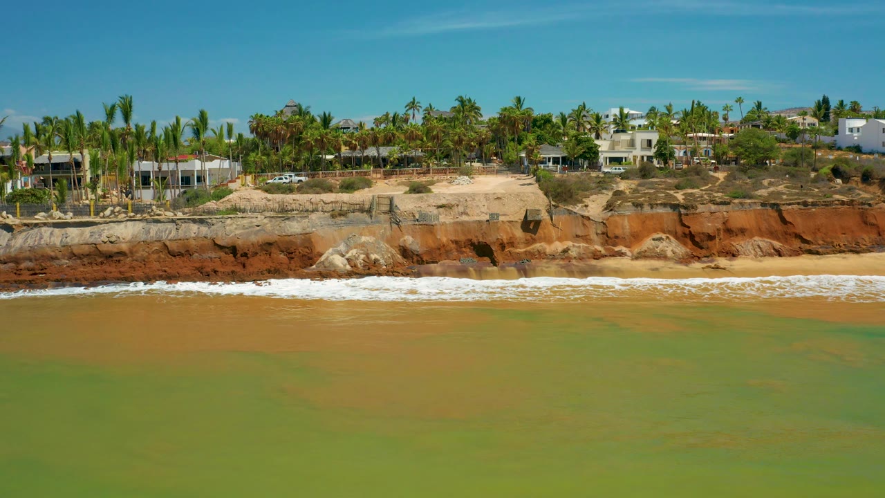 Ocean water and beach below orange cliffs with palms and buildings above.