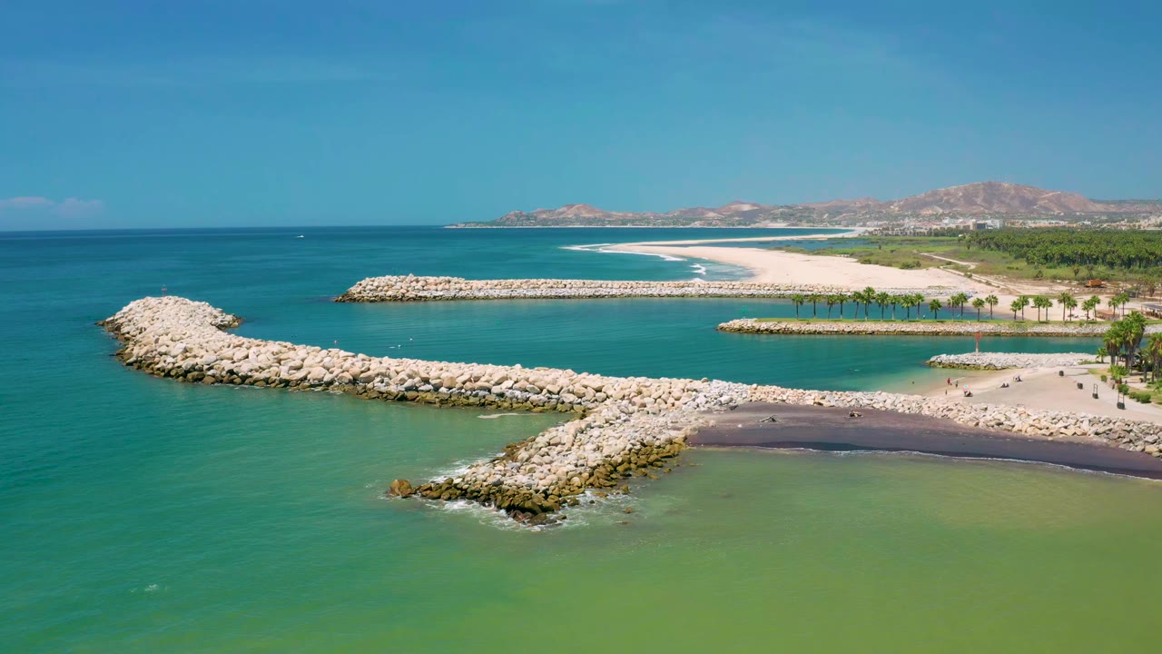 Aerial frame from San Jose Del Cabo, Mexico showing pale rock breakwaters crossing turquoise water toward a sandy beach, palm trees, distant hills, clear sky, and no visible people.