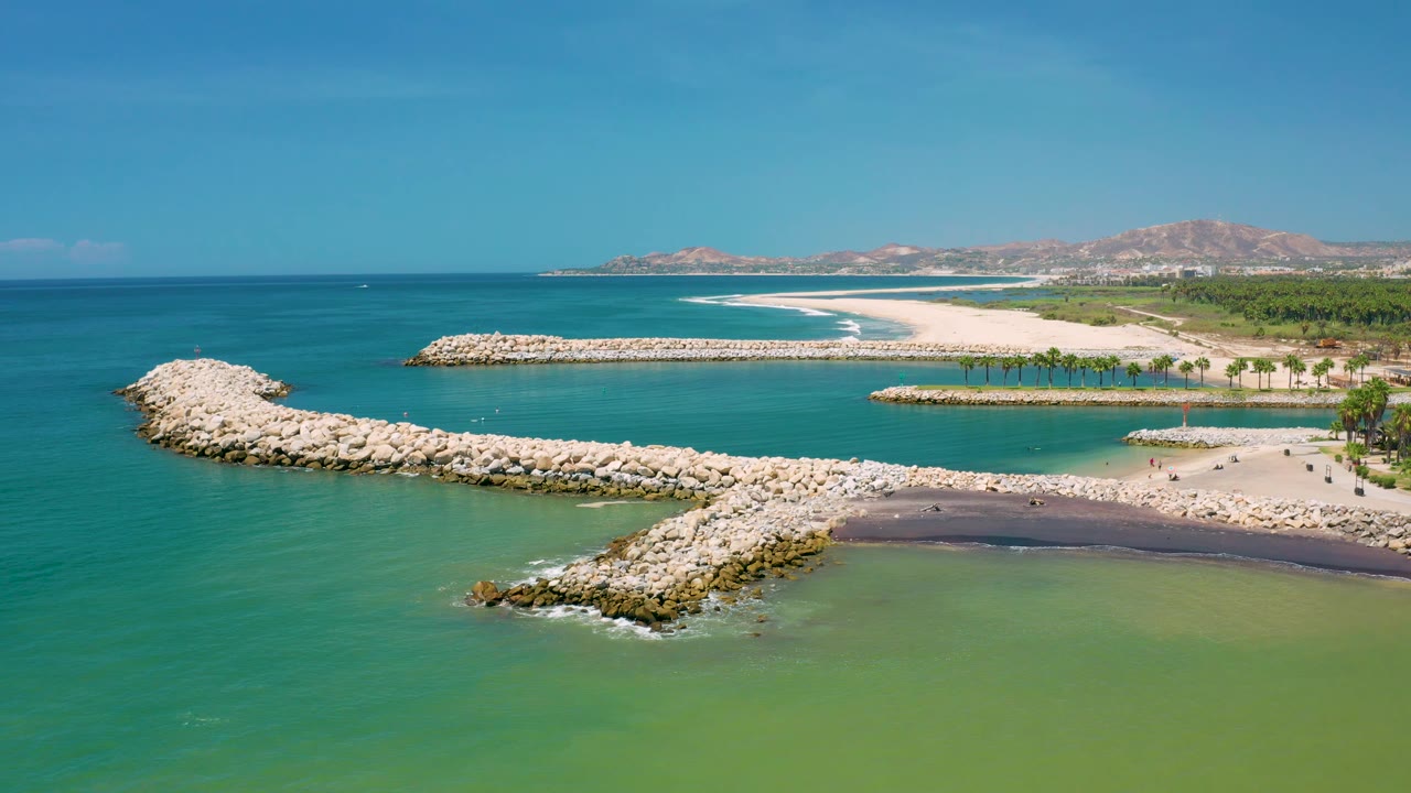 Aerial frame from San Jose Del Cabo, Mexico showing a long rock jetty angled across clear water, a second rock barrier offshore, a sandy shoreline, low hills, and no visible people.
