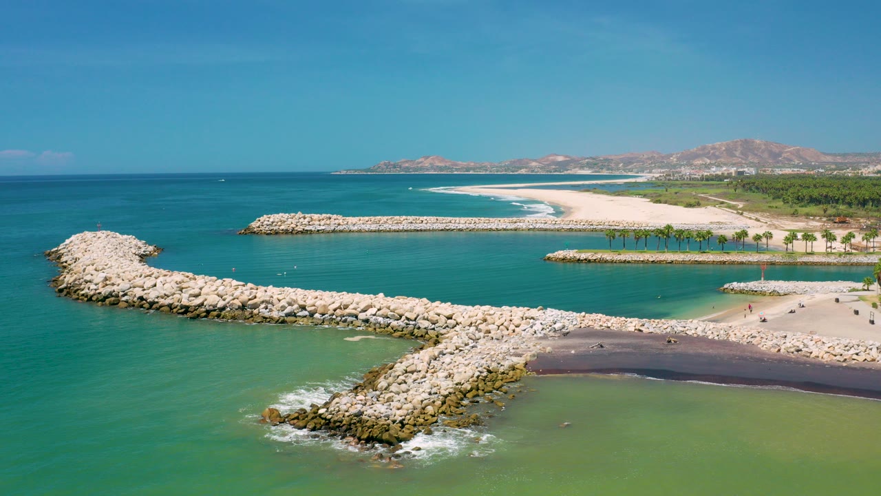 San Jose Del Cabo aerial frame with pale rock reef arms crossing green-blue water, a bright sandbar and palm-lined beach beyond, and no recognizable people visible.