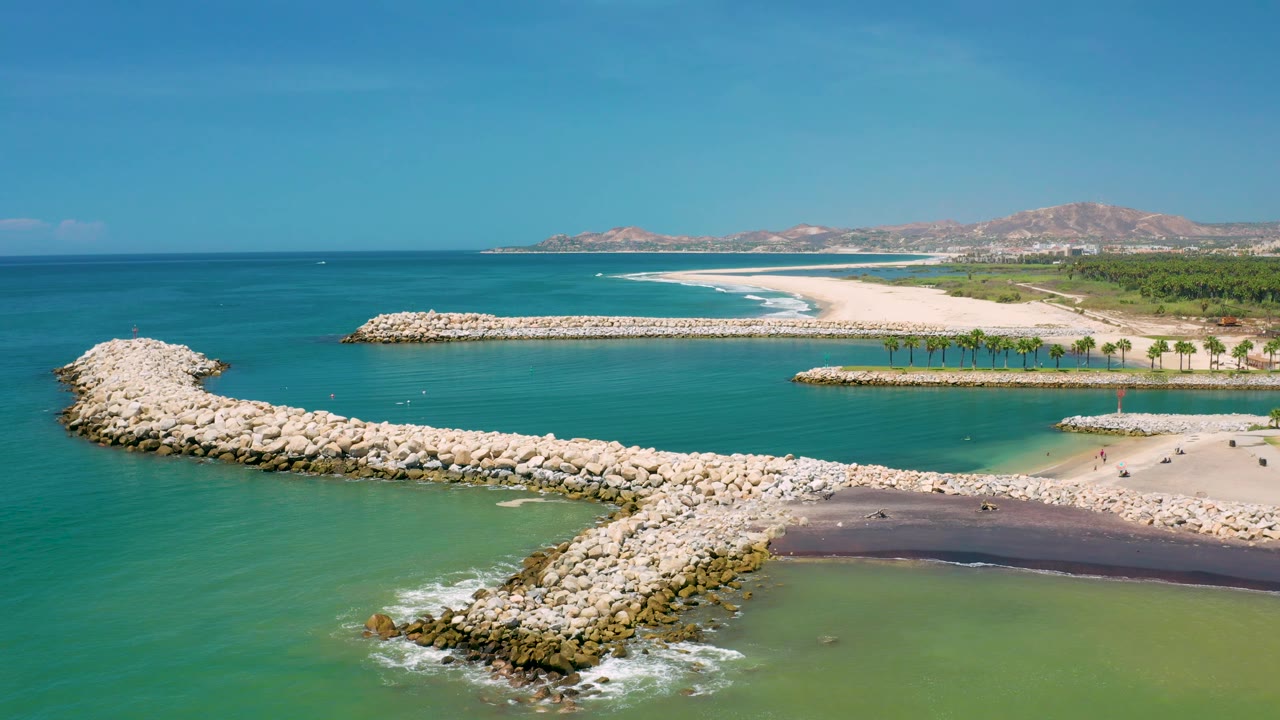 San Jose Del Cabo aerial frame with reef rocks in the foreground, green shallows at lower right, white sandbars in the midground, and no recognizable people visible.