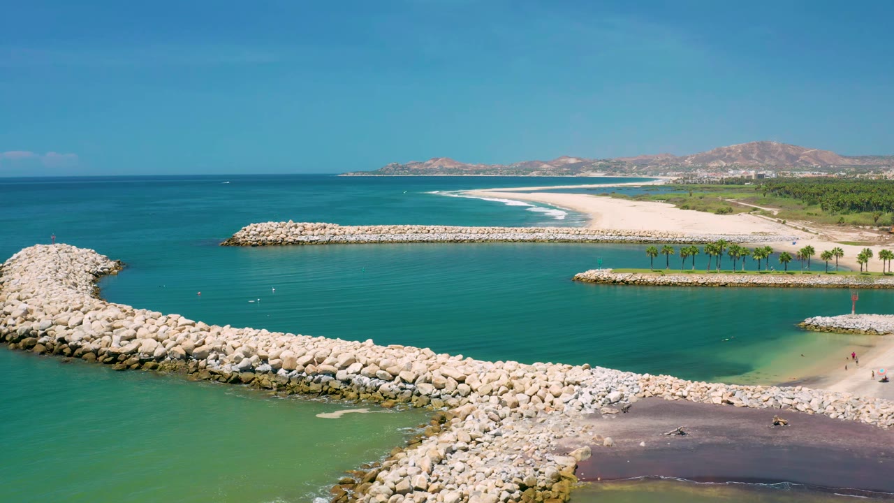 Aerial view in San Jose Del Cabo, Mexico with the pale rock breakwater crossing the lower frame, a calm inlet inside it, white surf lines on the beach, palm rows, and no clearly visible people.