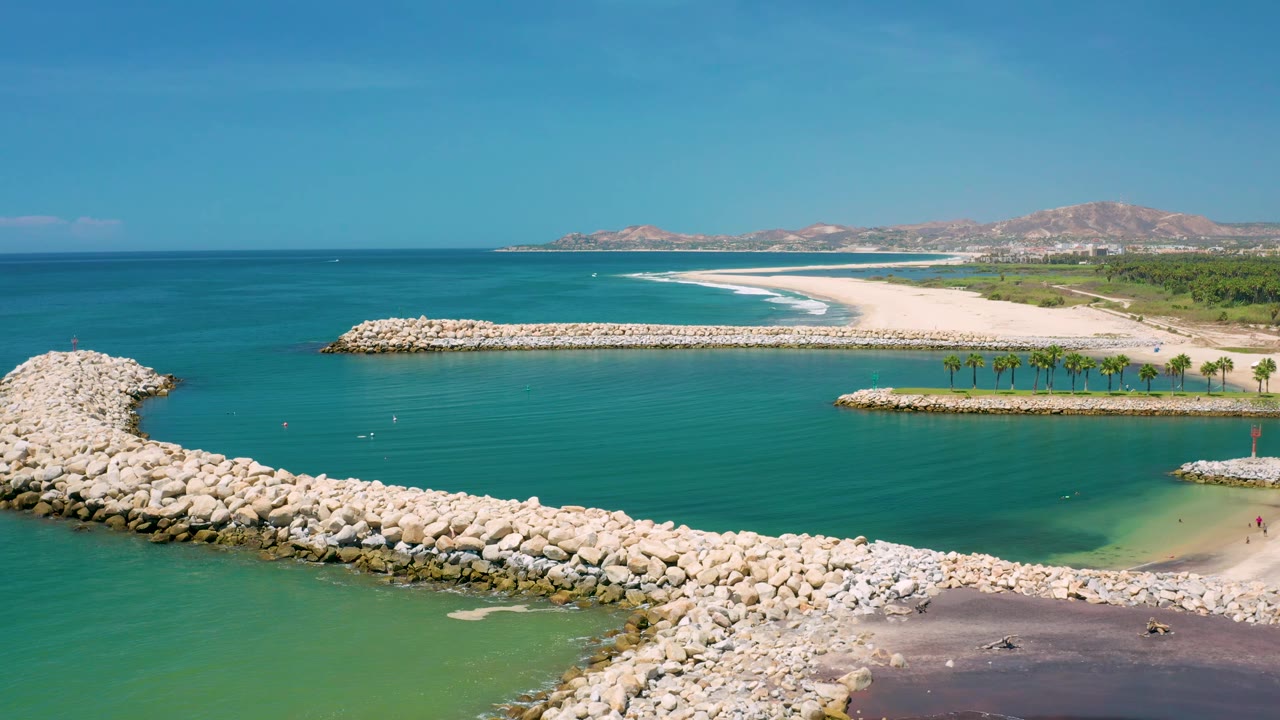 Wide 4K aerial frame in San Jose Del Cabo, Mexico showing open turquoise sea, the breakwater curving through the lower frame, a long sandy beach, low hills, and no clearly visible people.