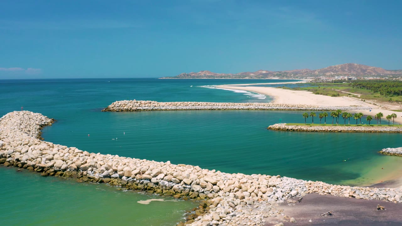 San Jose Del Cabo, Mexico aerial frame with breakwater rocks filling the lower left, calm teal water inside the structure, surf bands along the beach, hills on the horizon, and no clearly visible people.