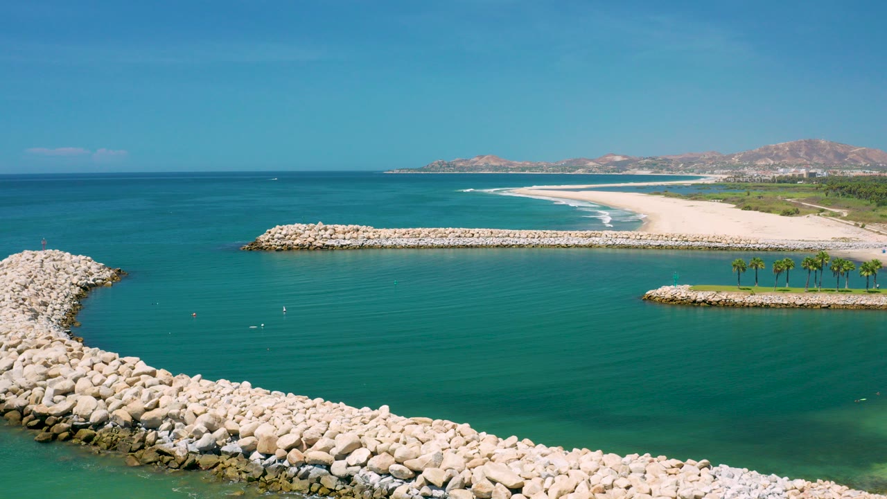 Aerial frame over San Jose Del Cabo harbor water, with pale rock seawalls in the foreground, a sandy beach and low mountains in the distance, and no visible people.