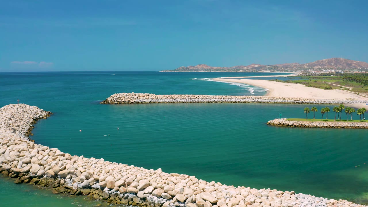 Aerial view in San Jose Del Cabo with a curved rock breakwater across the midground, calm turquoise water below, distant beach surf, palms, and no visible people.