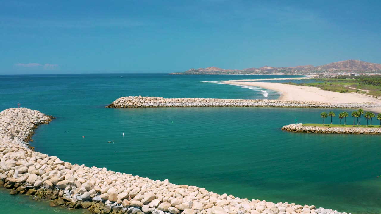 Wide aerial frame of San Jose Del Cabo showing two pale rock seawalls framing a small harbor opening, with teal water, beach surf, palms, mountains, and no visible people.