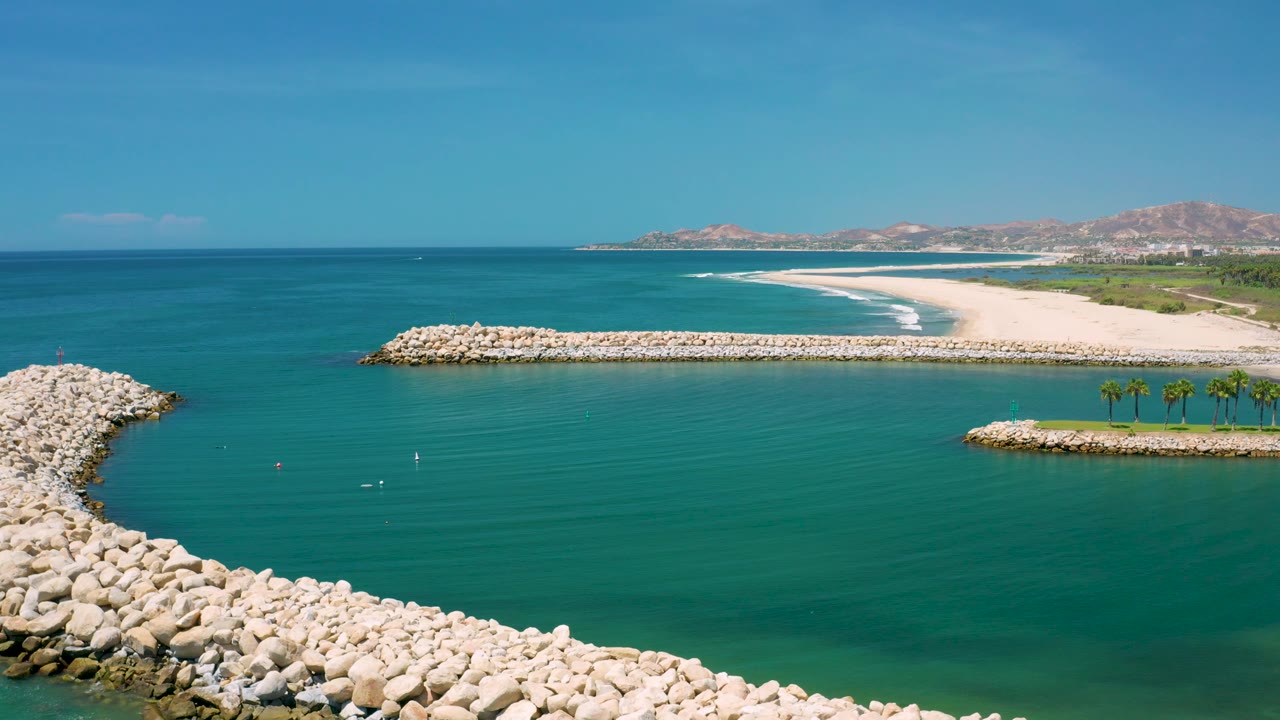 Aerial frame over San Jose Del Cabo harbor water, where foreground rocks border the lower edge and a long sandy beach with small surf lines stretches behind, with no visible people.