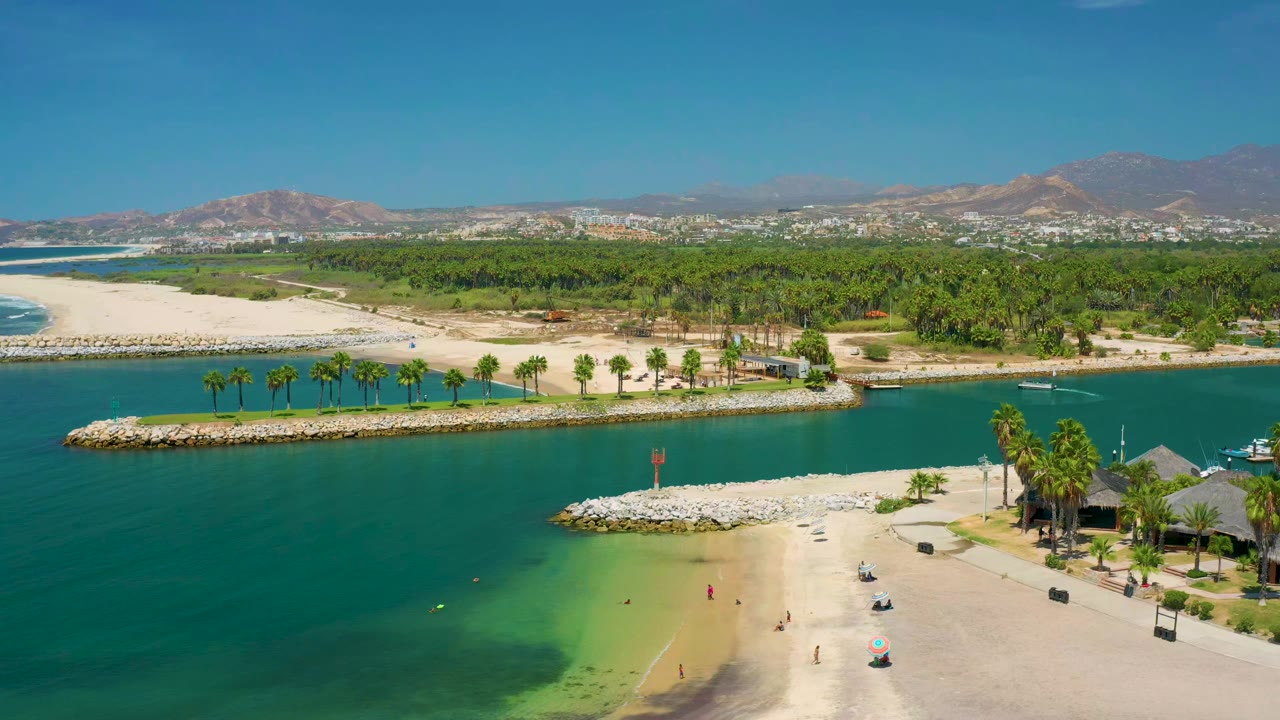 High aerial view of a beach cove, rock jetties, palms, green parkland, and mountains.