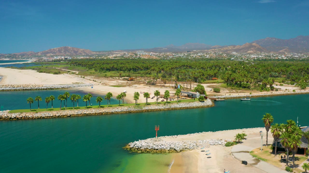 Aerial frame from San Jose Del Cabo shows green canal water below sandy banks, a palm-lined edge, a broad grove, distant buildings, brown mountains, and no visible people.