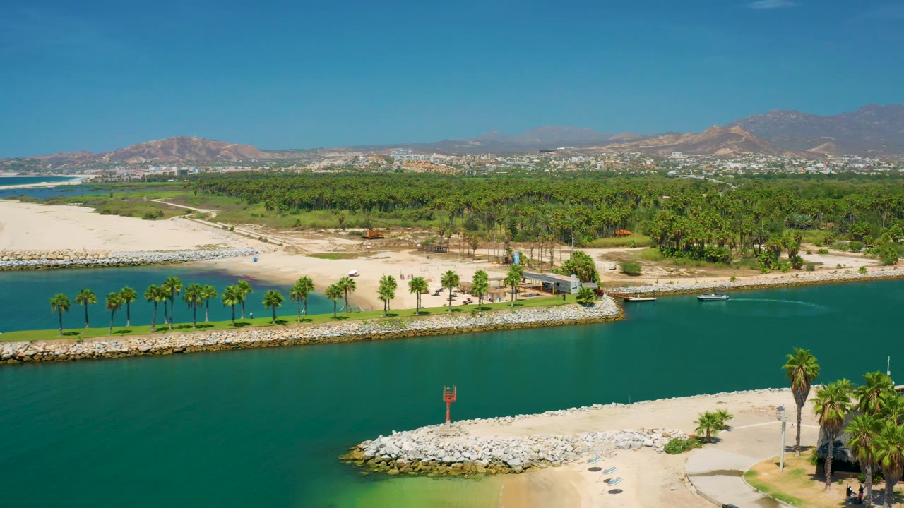 Aerial view in San Jose Del Cabo shows a palm-lined canal edge, pale sand, rock breakwater, small boats near shore, distant development, mountains, and no visible people.