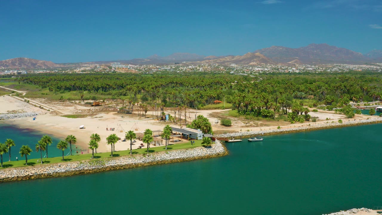 Wide aerial frame over San Jose Del Cabo, Mexico, with blue-green water in front, a rock-edged palm shoreline, pale beach, dense trees, distant city buildings, and mountains beyond.