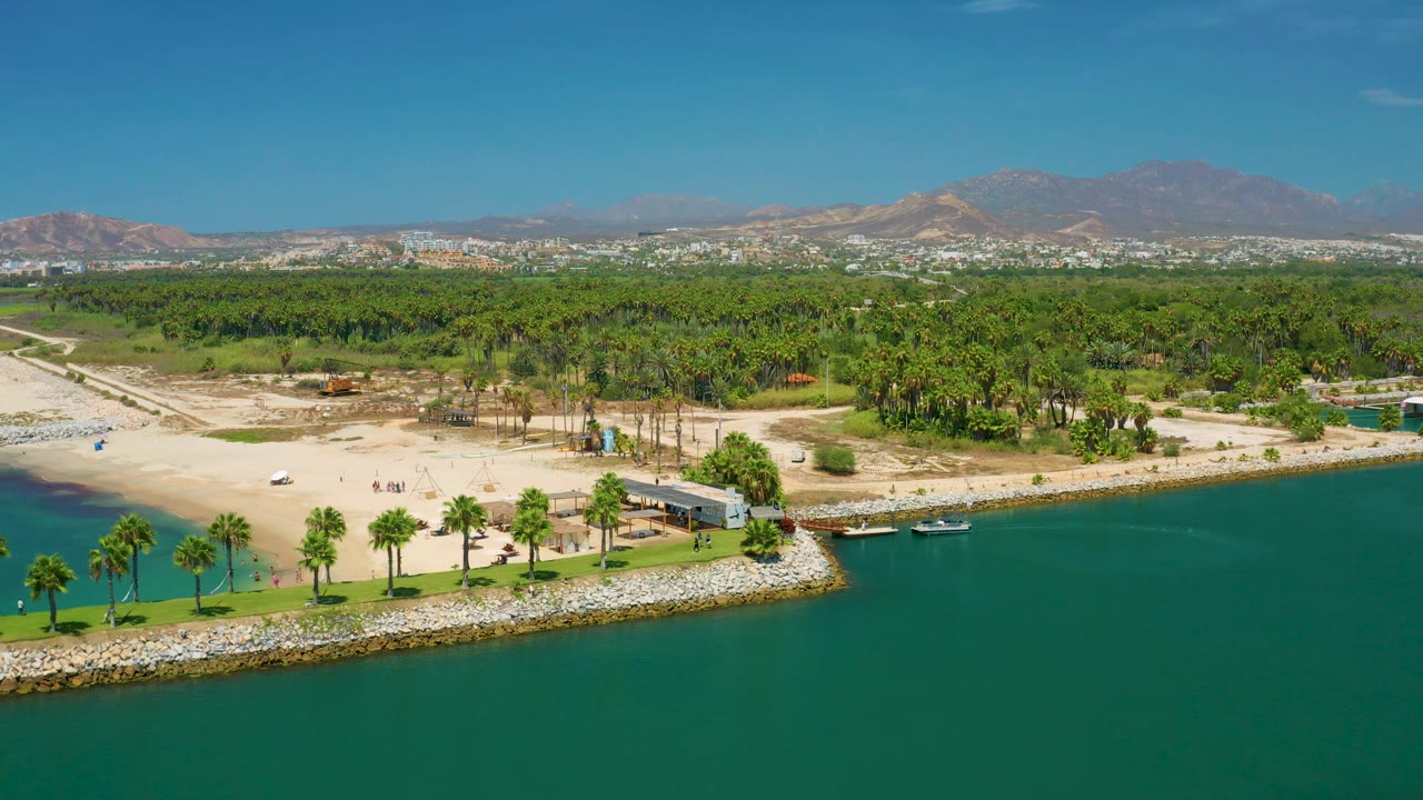 San Jose Del Cabo aerial frame with broad blue-green water at the bottom, a palm-lined strip of shore, pale sand, thick greenery, distant city blocks, and mountain ridges.